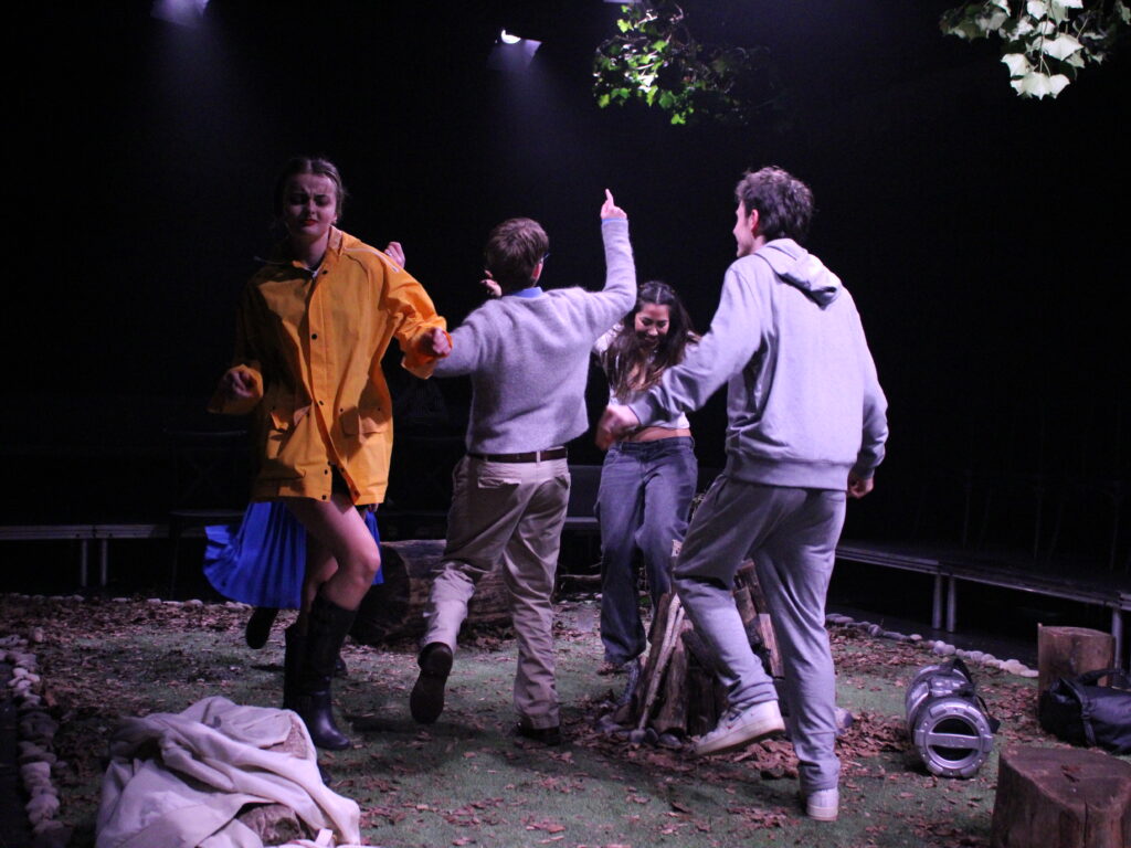 Five people move energetically around a central campfire structure on a stage dressed as a woodland clearing. One person in a yellow coat lifts a leg mid‑step, while others reach upward or lean into the movement. Leaves, branches and scattered props cover the ground under overhead stage lighting.