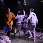 Five people move energetically around a central campfire structure on a stage dressed as a woodland clearing. One person in a yellow coat lifts a leg mid‑step, while others reach upward or lean into the movement. Leaves, branches and scattered props cover the ground under overhead stage lighting.