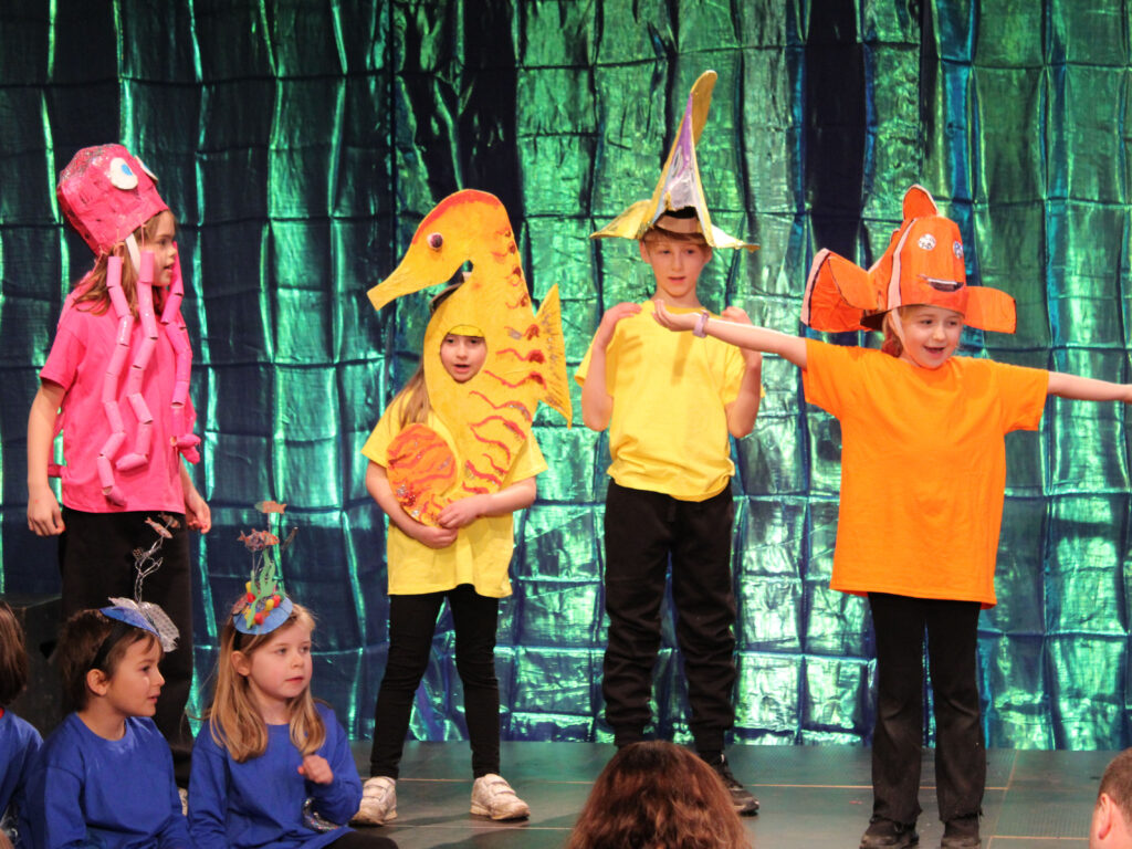 Four children stand on a school hall stage dressed in black trousers and brightly-coloured t-shirts, with an iridescent backdrop. One in pink wears an octopus headdress. another in yellow is dressed as a seahorse. Two others wear fish headdresses, one yellow and another orange. Two children sit on the floor of the stage, dressed in blue.