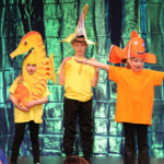 Four children stand on a school hall stage dressed in black trousers and brightly-coloured t-shirts, with an iridescent backdrop. One in pink wears an octopus headdress. another in yellow is dressed as a seahorse. Two others wear fish headdresses, one yellow and another orange. Two children sit on the floor of the stage, dressed in blue.