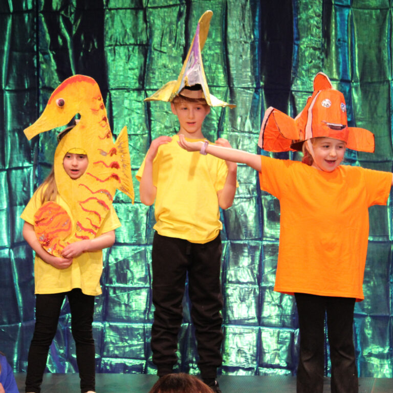 Four children stand on a school hall stage dressed in black trousers and brightly-coloured t-shirts, with an iridescent backdrop. One in pink wears an octopus headdress. another in yellow is dressed as a seahorse. Two others wear fish headdresses, one yellow and another orange. Two children sit on the floor of the stage, dressed in blue.