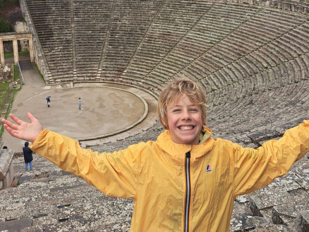 Smiling boy in a yellow jacket posing on an ancient stone amphitheater with forested hills behind him.