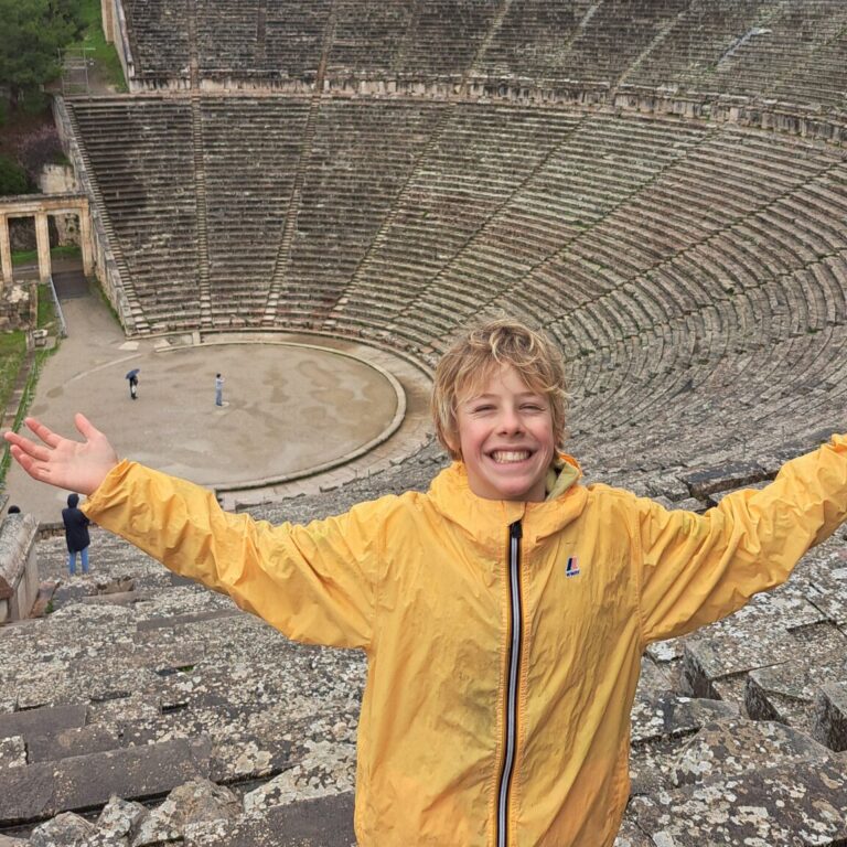 Smiling boy in a yellow jacket posing on an ancient stone amphitheater with forested hills behind him.