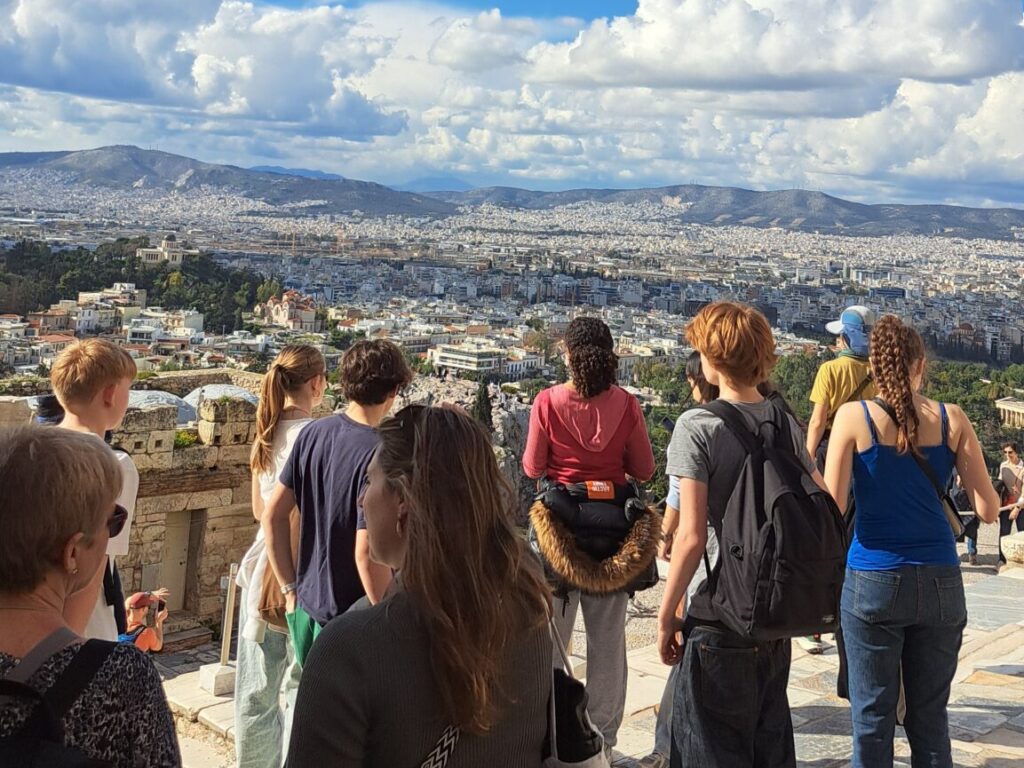A group of students stand at a stone viewpoint overlooking a city spread across a valley with hills in the distance under a blue, cloudy sky.