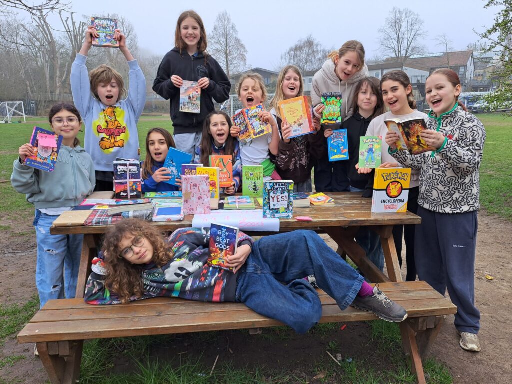 Group of children outdoors around a picnic table, holding colorful books and smiling at the camera.
