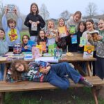 Group of children outdoors around a picnic table, holding colorful books and smiling at the camera.