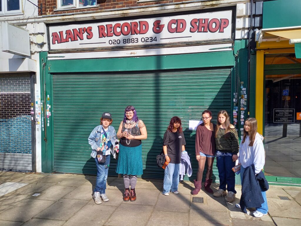 Group of seven young people posing in front of a closed music shop with a green rolling shutter and 'Alan's Record & CD Shop' sign above them.