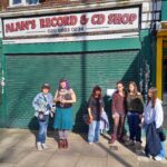 Group of seven young people posing in front of a closed music shop with a green rolling shutter and 'Alan's Record & CD Shop' sign above them.