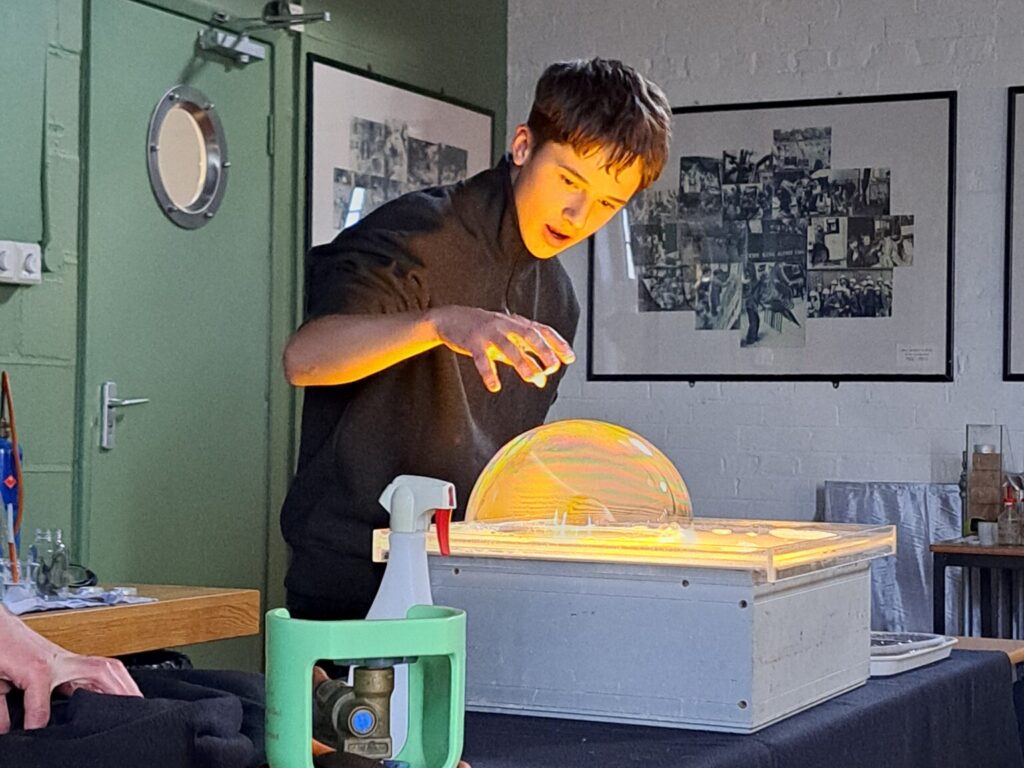 Male student stands above a projector, looking at a huge bubble on its surface. His face is lit by the projector's light, his face showing an expression of wonder, and his hand hovers above the bubble.