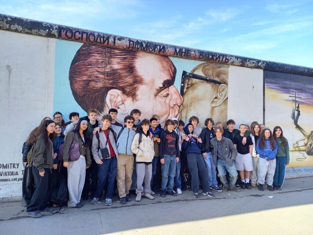 Group of students posing in front of a large wall mural (part of the Berlin Wall) showing two close-up faces nearby each other, with the group at the bottom of the image to the left and center.