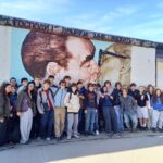 Group of students posing in front of a large wall mural (part of the Berlin Wall) showing two close-up faces nearby each other, with the group at the bottom of the image to the left and center.