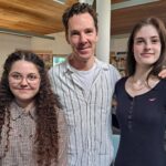 Three people stand together indoors in a library, smiling for the camera; a man in the middle flanked by two young women. The man is actor Benedict Cumberbatch.