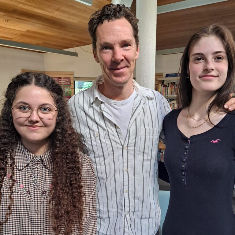 Three people stand together indoors in a library, smiling for the camera; a man in the middle flanked by two young women. The man is actor Benedict Cumberbatch.