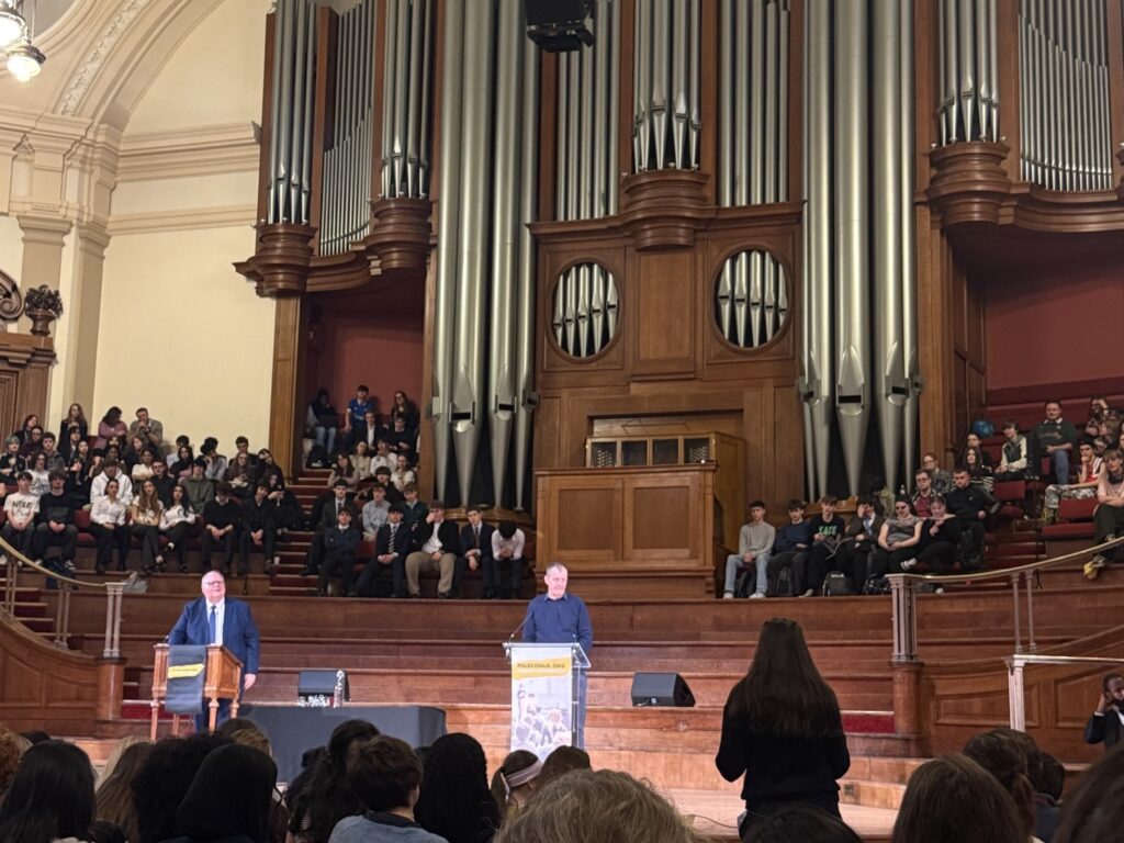 Alastair Campbell standing at a lectern on stage in a grand hall, with an impressive organ dramatically lit behind him. In the foreground, an audience can be seen in shadow, with one standing to ask a question.