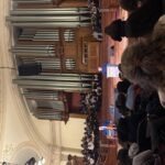 Alastair Campbell standing at a lectern on stage in a grand hall, with an impressive organ dramatically lit behind him. In the foreground, an audience can be seen in shadow, with one standing to ask a question.