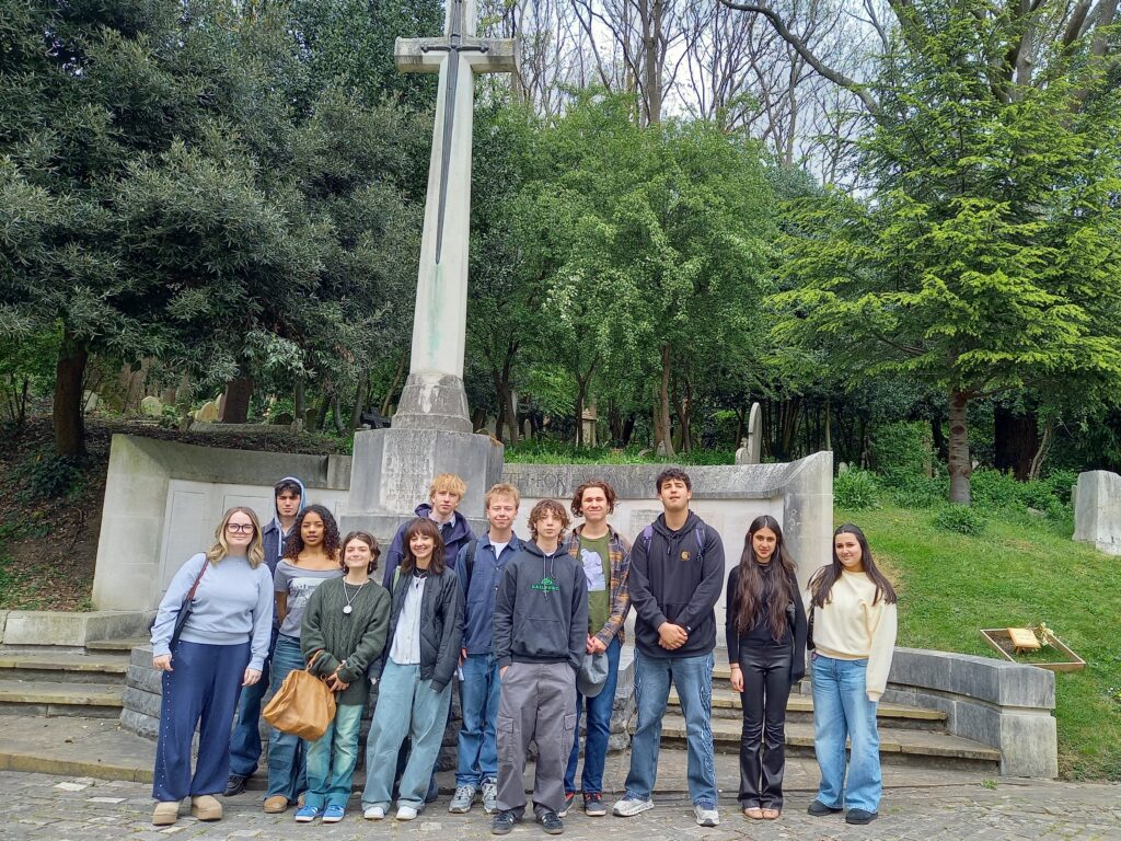Group of young people posing on steps in a cemetery with a tall cross monument and green trees behind them.
