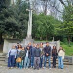 Group of young people posing on steps in a cemetery with a tall cross monument and green trees behind them.