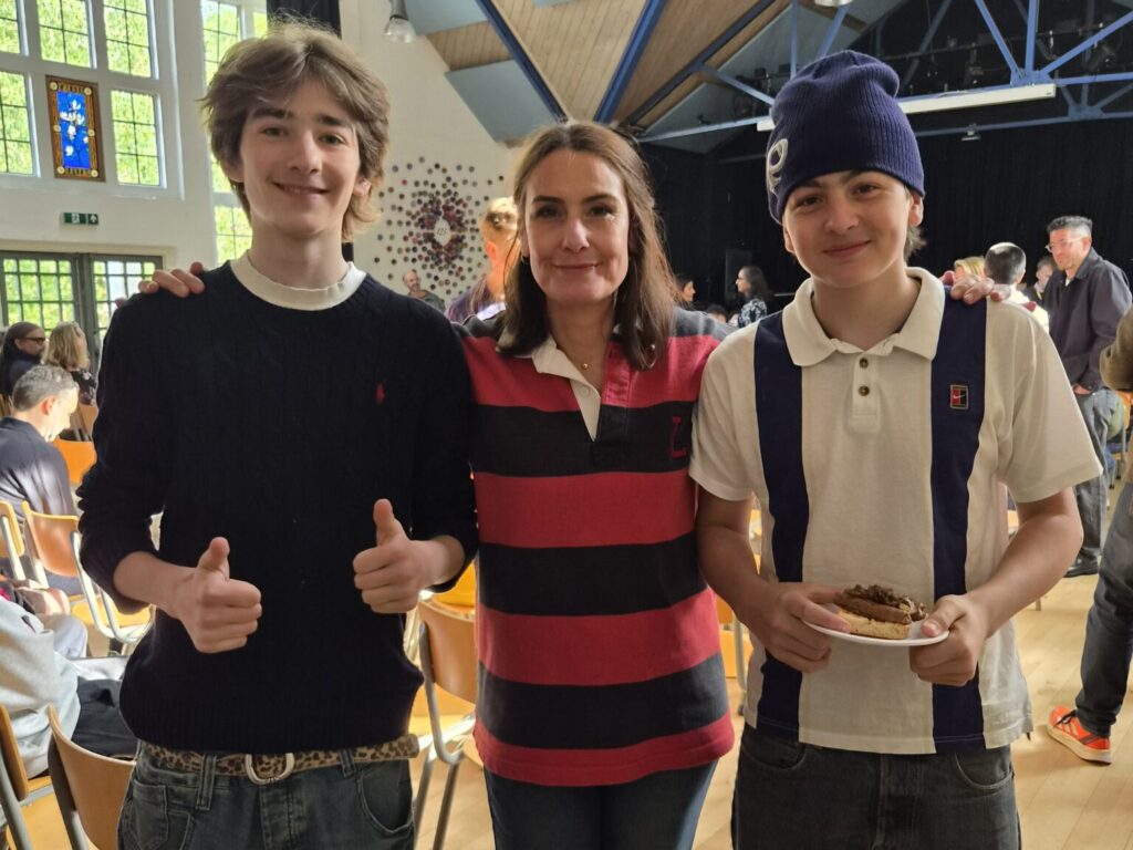 Three smiling people posing together for a photo in a bright hall with wooden floor and chairs in the background, left and center are teens/adult, right a young boy holding a plate with cake or pastry.