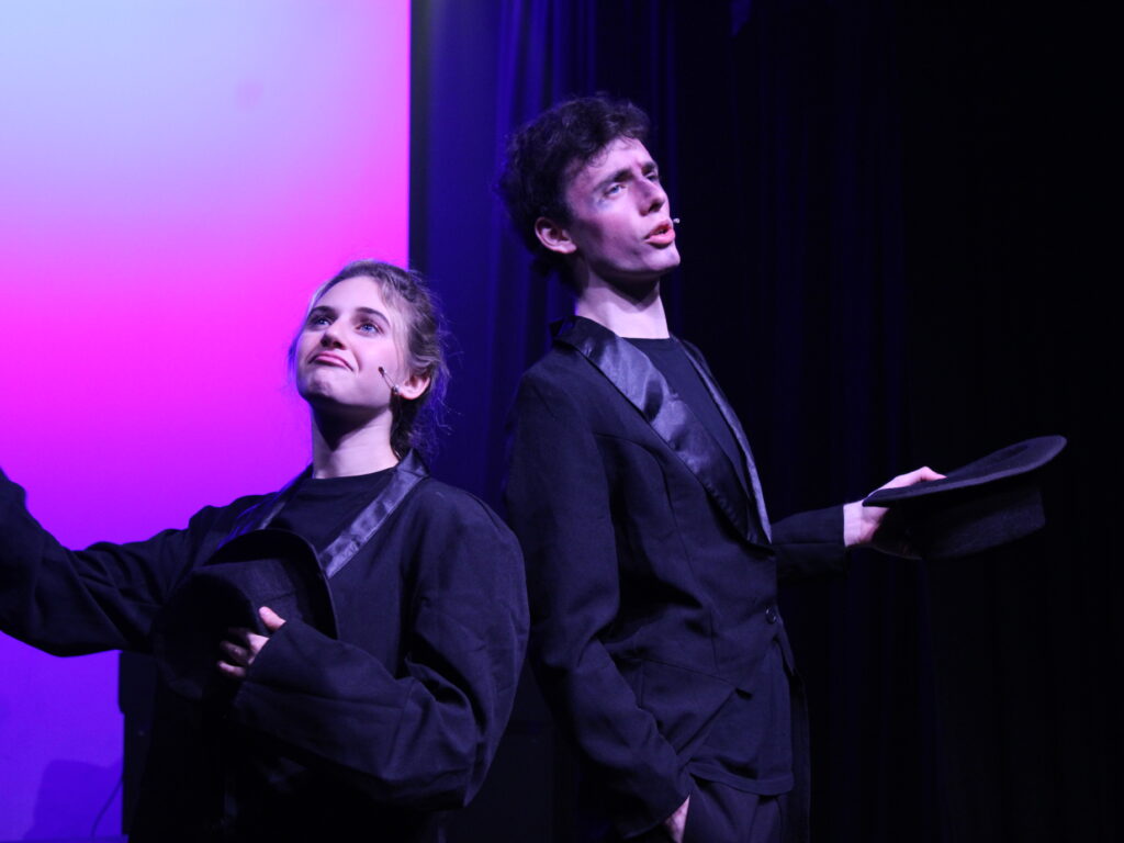 Two performers in black stage costumes pose on a purple-lit stage, one holding a black hat.