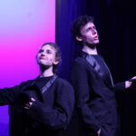 Two performers in black stage costumes pose on a purple-lit stage, one holding a black hat.