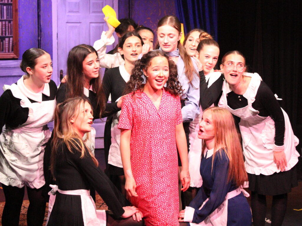 Group of young performers in black-and-white maid costumes posing together on a theater stage with a purple backdrop and wooden set pieces behind them.