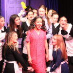 Group of young performers in black-and-white maid costumes posing together on a theater stage with a purple backdrop and wooden set pieces behind them.