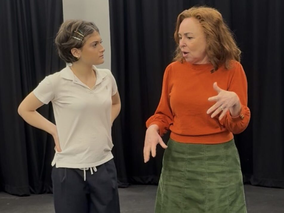 Two women stand on a theatre stage in conversation; one in an orange sweater and green skirt, the other in a white polo and dark pants, with black curtains behind them.