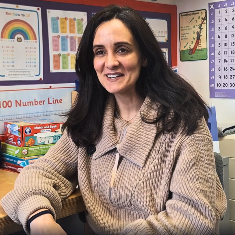 Smiling woman in a beige sweater seated at a classroom desk with math posters on the wall behind her.