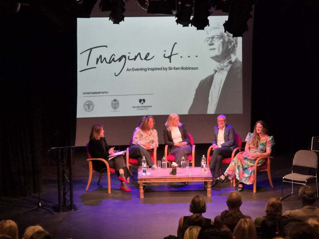 Five-panel discussion on a stage with red chairs, audience in foreground; a large screen behind shows 'Imagine if...' and a portrait of Sir Ken Robinson.