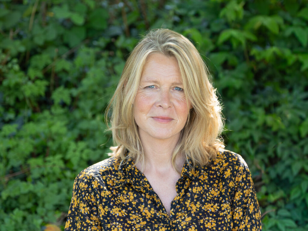 Portrait of a woman with blonde hair outdoors, wearing a black dress with yellow flowers, greenery in the background.