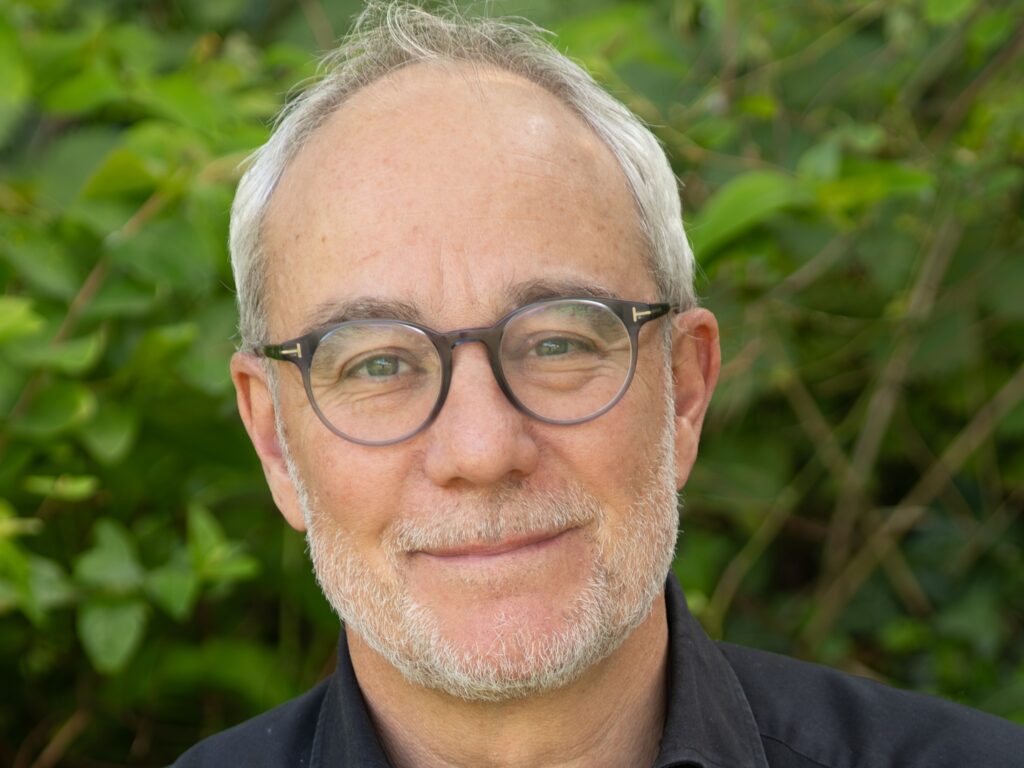 Portrait of a middle‑aged man with gray hair and round glasses, outdoors in greenery, wearing a black shirt with a subtle smile.