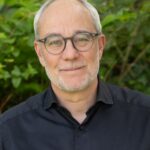 Portrait of a middle‑aged man with gray hair and round glasses, outdoors in greenery, wearing a black shirt with a subtle smile.