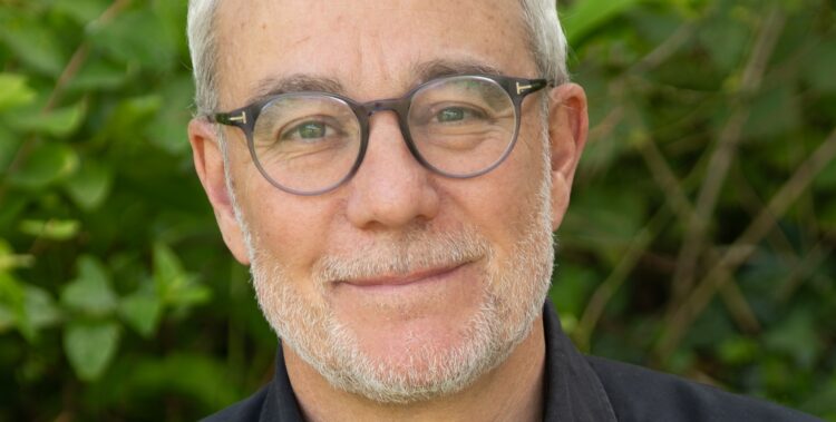 Portrait of a middle‑aged man with gray hair and round glasses, outdoors in greenery, wearing a black shirt with a subtle smile.