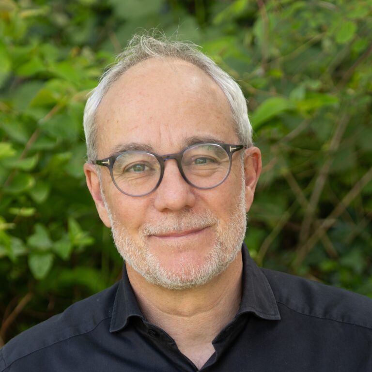 Portrait of a middle‑aged man with gray hair and round glasses, outdoors in greenery, wearing a black shirt with a subtle smile.