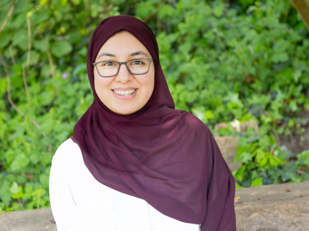 Smiling woman wearing a maroon hijab and glasses, outdoors with green foliage in the background.