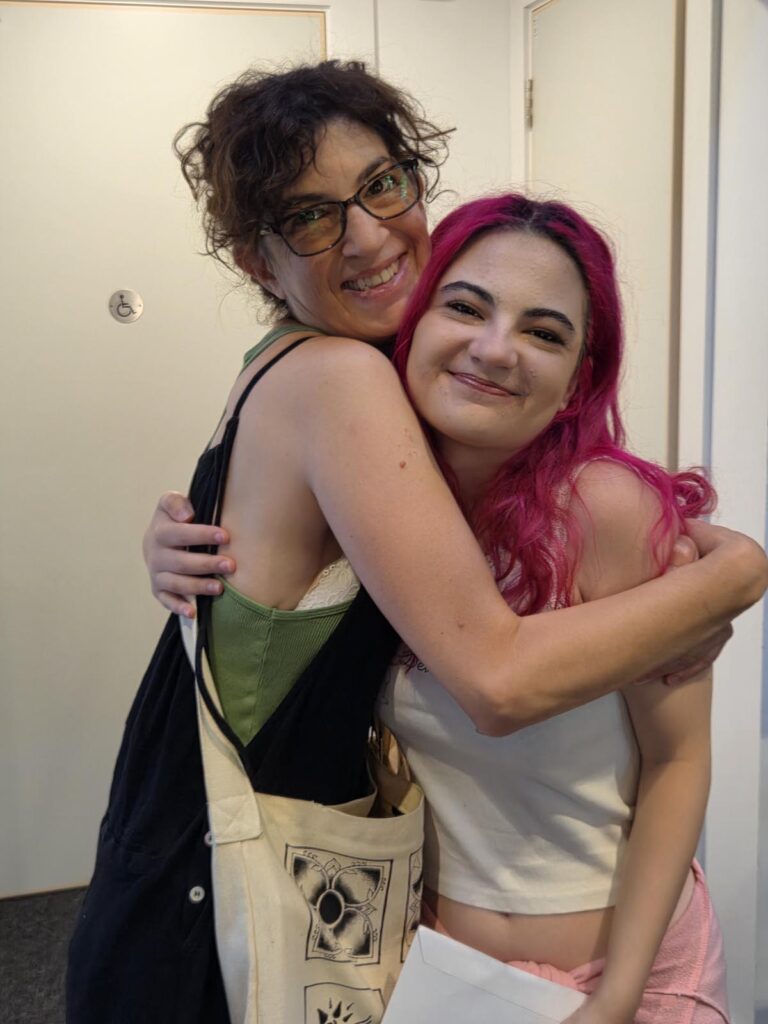 Two women smiling and hugging each other in a doorway, one with pink hair and the other with short brown hair and glasses, both wearing casual outfits.