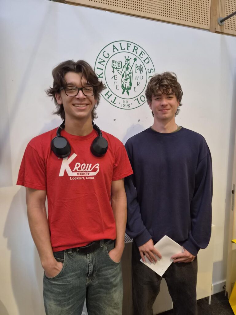Two young men pose for a photo indoors in front of a crest logo; one wears a red t-shirt with headphones, the other a dark sweatshirt holding papers.