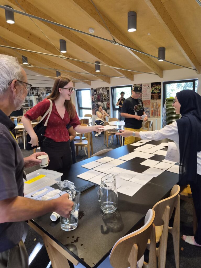 People standing around a long black table covered with white cards in a bright room with wooden beams, participating in a workshop or sign-in activity.