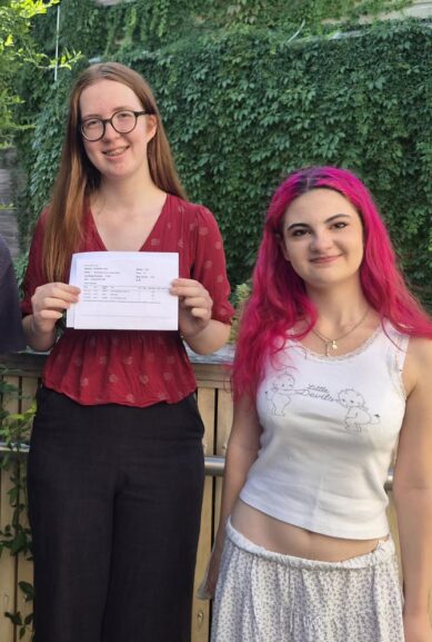 Three people pose outdoors by a wooden fence; center person shows a document, smiling at the camera.