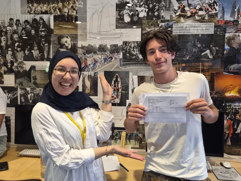Two smiling people pose in front of a collage wall; a woman wearing a hijab and staff lanyard on the left, a young man holding a document on the right.