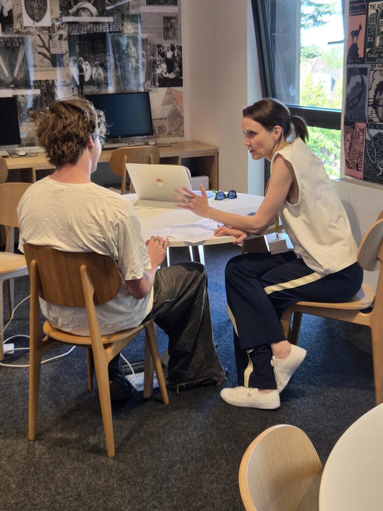 Tutor explains documents to a student across a table in a study room, laptop open nearby.