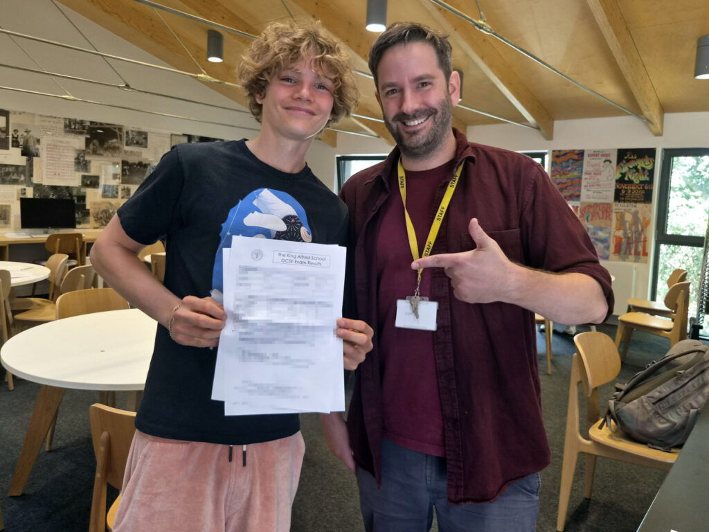 Smiling student holds GCSE results with a staff member giving a thumbs-up in a classroom.