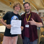 Smiling student holds GCSE results with a staff member giving a thumbs-up in a classroom.