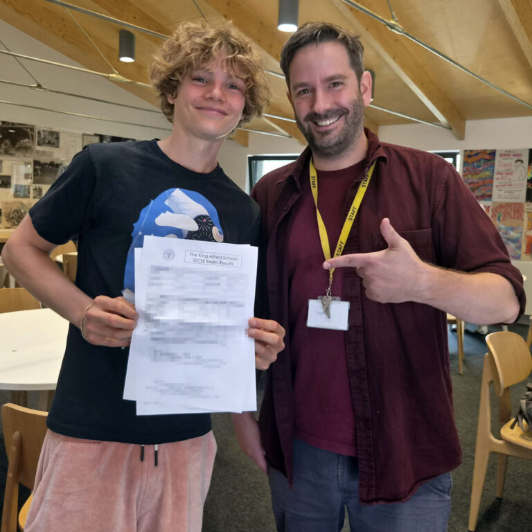 Smiling student holds GCSE results with a staff member giving a thumbs-up in a classroom.