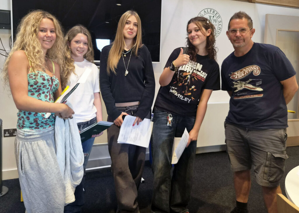 Group of five people posing indoors, four students holding papers and a man in a dark T-shirt, smiling at the camera in a classroom setting.