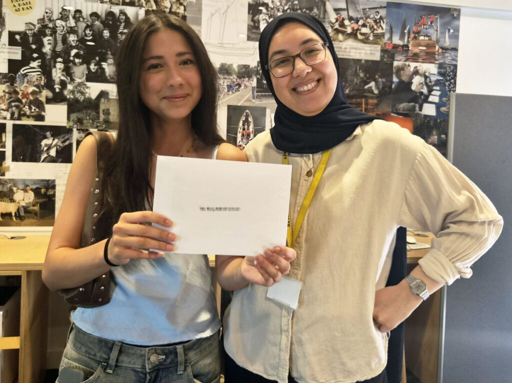 Two women smiling at the camera, holding a white document in a bright, collage-covered office background.