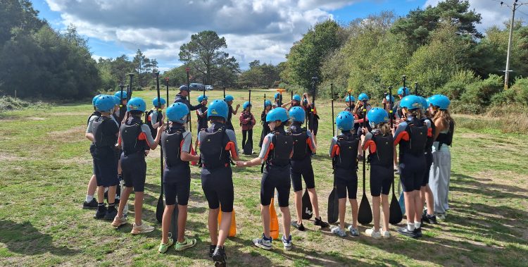 Large group of students wearing wetsuits and protective helmets hold oars and stand in a circle on some grass