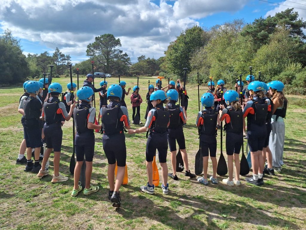 Large group of students wearing wetsuits and protective helmets hold oars and stand in a circle on some grass
