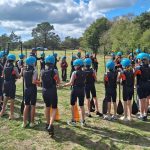 Large group of students wearing wetsuits and protective helmets hold oars and stand in a circle on some grass