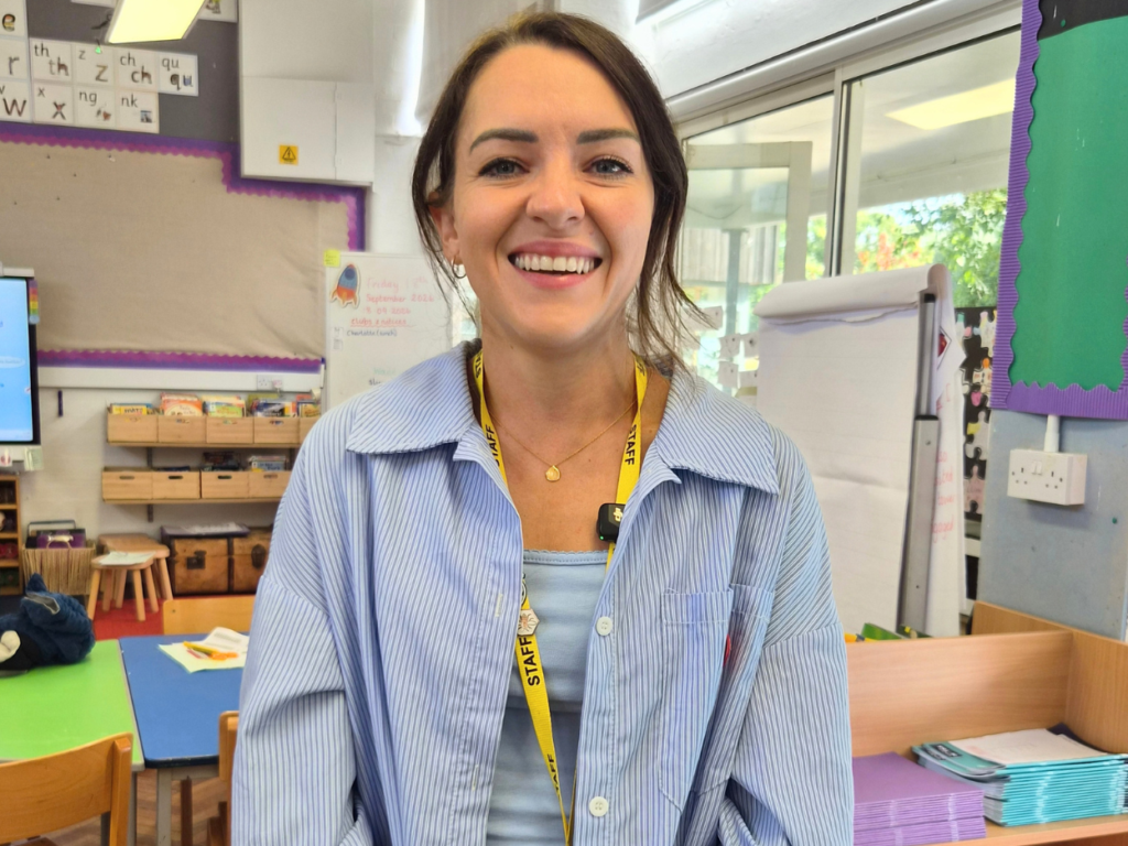 Headshot of a white female teacher, standing in a bright and colourful children's classroom. She has dark hair and is wearing a blue shirt with a yellow lanyard.